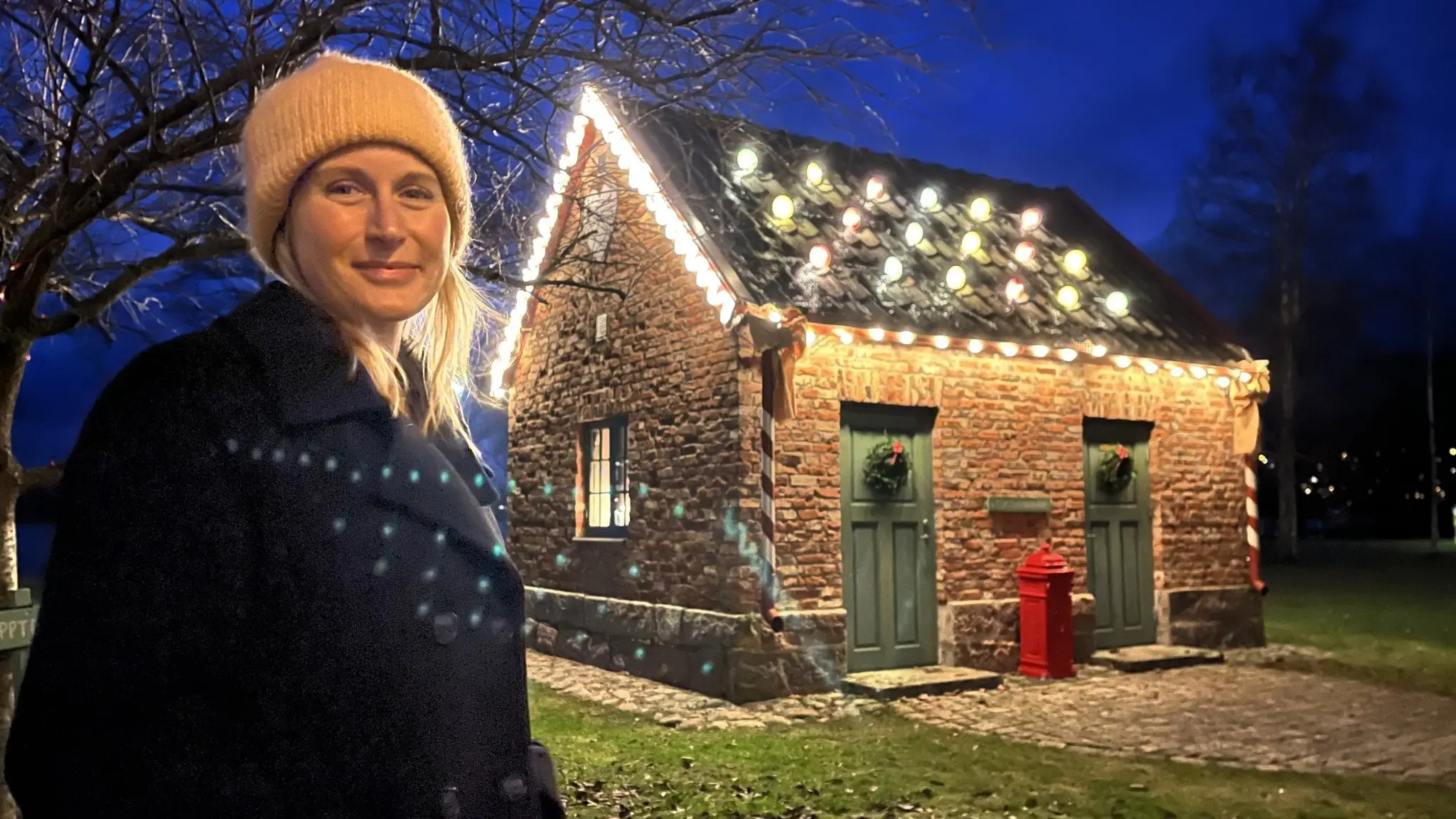 Ljusdesigner Mia Wengbrand står framför en liten stenbyggnad dekorerad med julbelysning, omgiven av träd och gräs i skymningsljus.