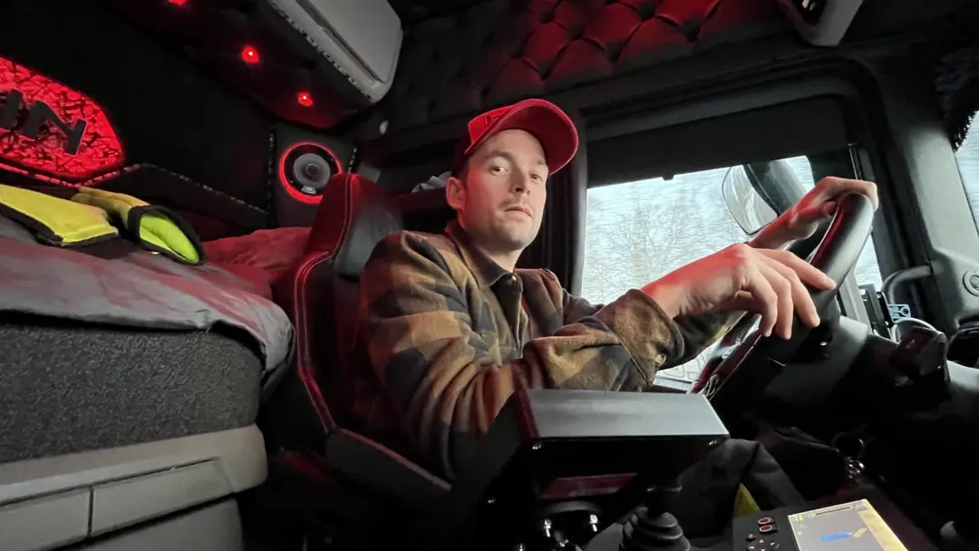 En person i arbetskläder sitter i förarsätet på en saltbil och håller i ratten, omgiven av en upplyst interiör. Utanför fönstret syns snö på marken.