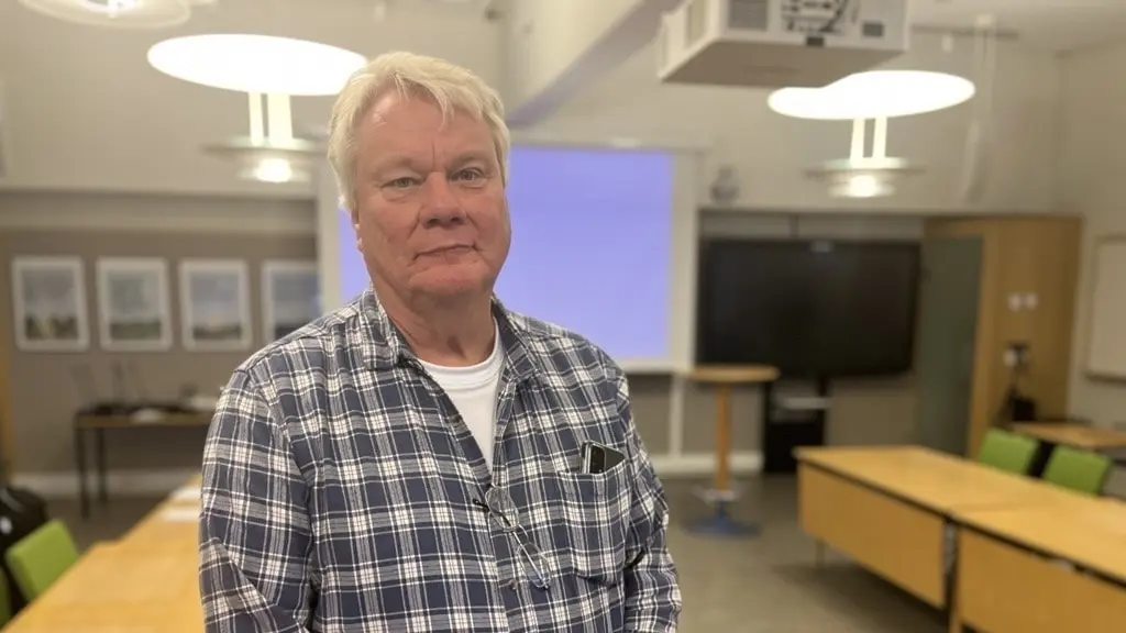 Håkan Cronelid står i ett konferensrum med projektor, vita väggar och gröna stolar i bakgrunden. Han har på sig en blå- och vitrutig skjorta.