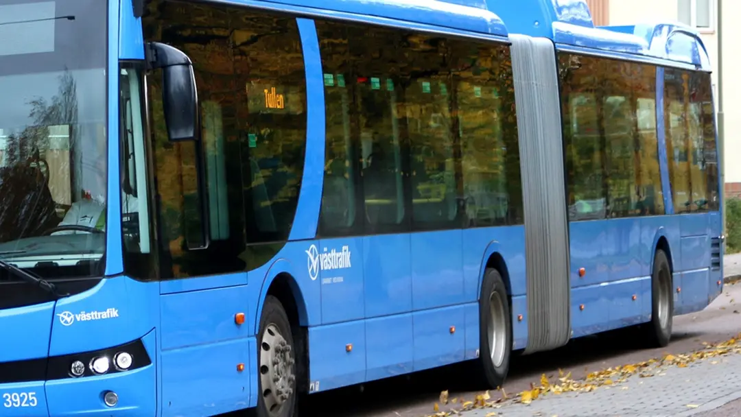 En blå Västtrafik-buss står parkerad vid en trottoar på en höstklädd gata. Bussens logotyp och namn syns tydligt på sidan.