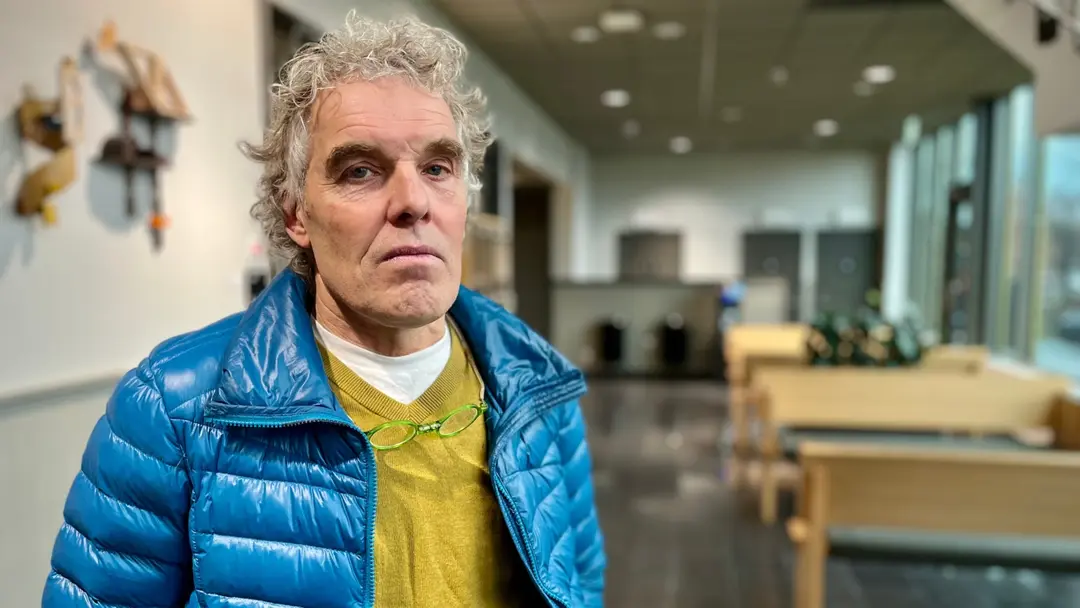 Per Brinkberg, klädd i blå jacka och gul tröja, står i en ljus och modern miljö med stora fönster och sittgrupper i bakgrunden. Bilden ger ett intryck av en offentlig plats eller kontorsmiljö.