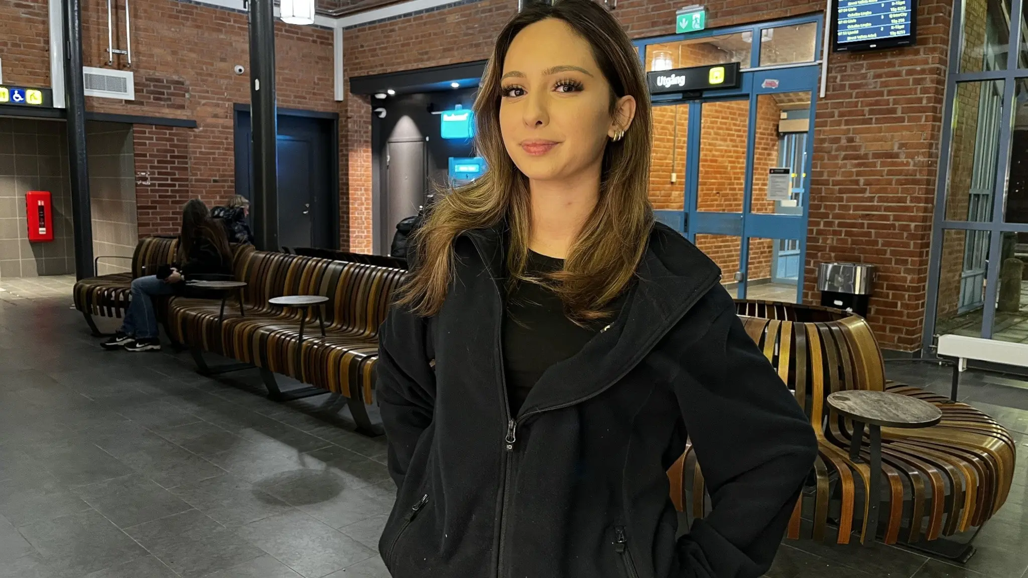 Amanda Warner står inomhus i en vänthall med tegelväggar, sittplatser och dörrar i bakgrunden. Hon bär en svart jacka.