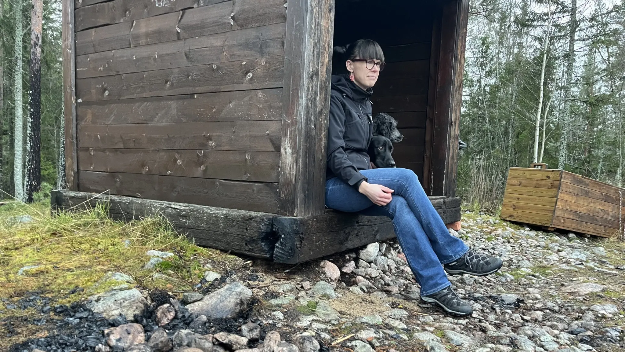 Elin Woxlin sitter vid ett vindskydd av trä i en skogsmiljö. Marken runtom är täckt av grus och brännmärken syns på vindskyddets bas efter en brand.