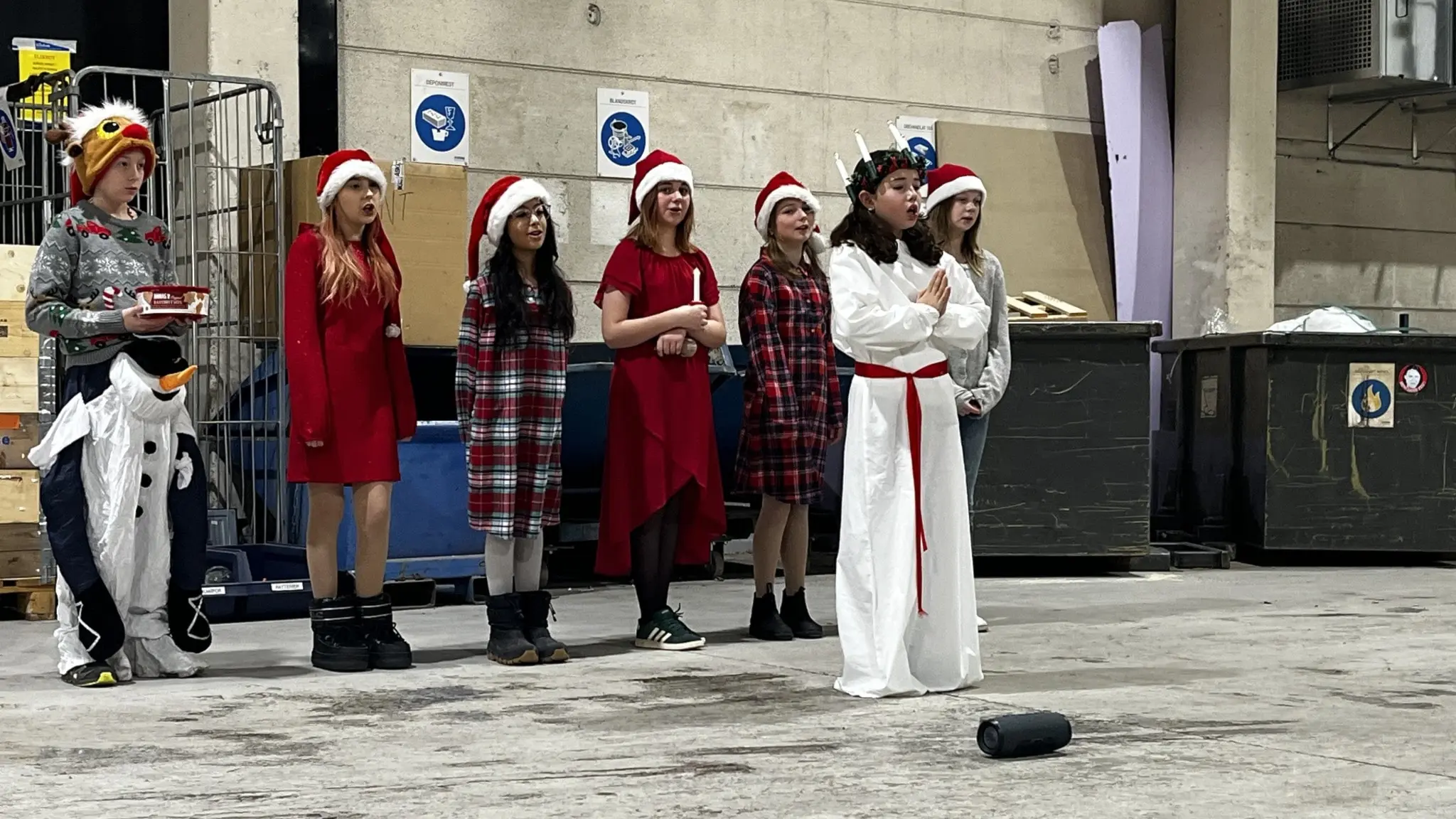 Femteklassare i Kilafors klädda i tomteluvor och luciakläder framför luciatåg i en industriell miljö, sprider julstämning och samlar in pengar till en klassresa.