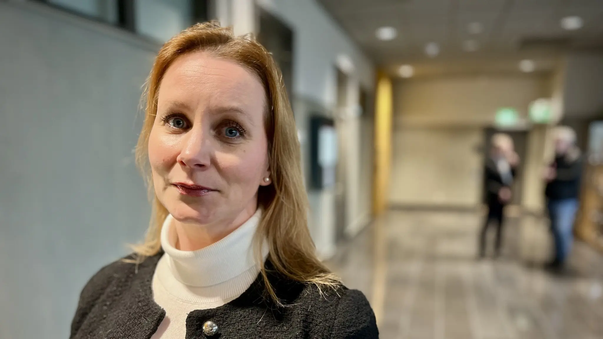 Nina Tiberg, advokat, står i en modern kontorsmiljö med suddig bakgrund som visar en korridor och människor i rörelse.