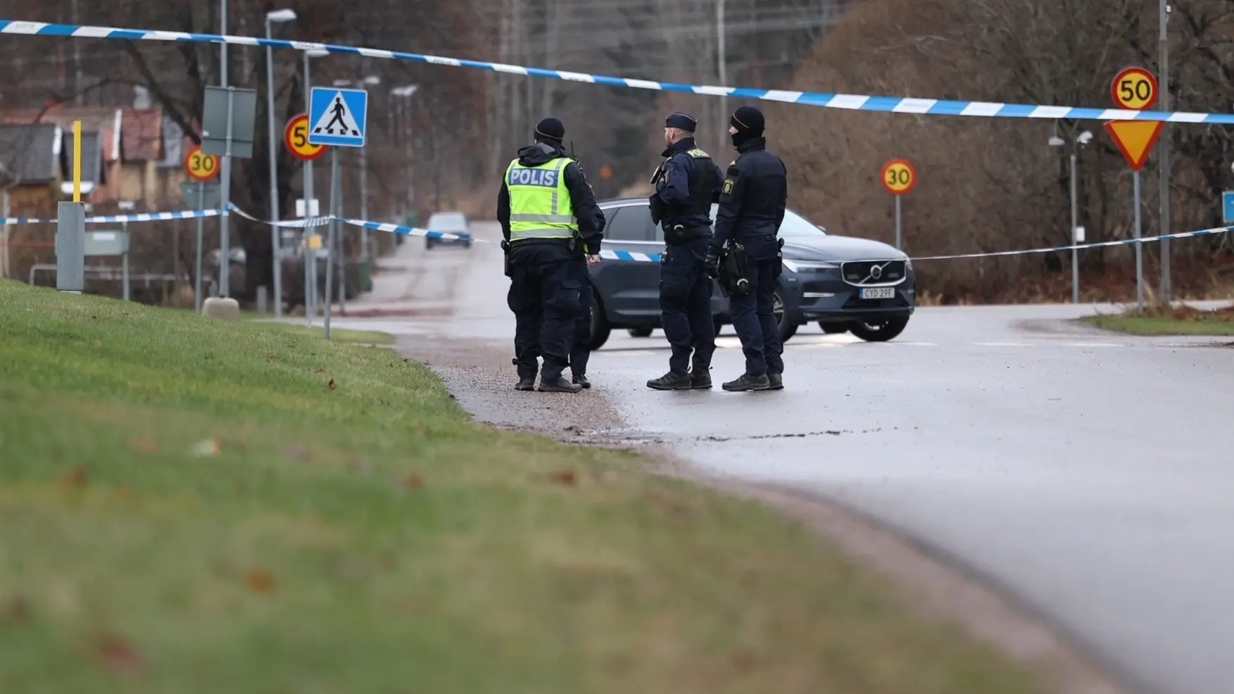 En avspärrad väg i ett bostadsområde med poliser på plats. De står vid en blåvit avspärrning, och en polisbil syns längre bort.