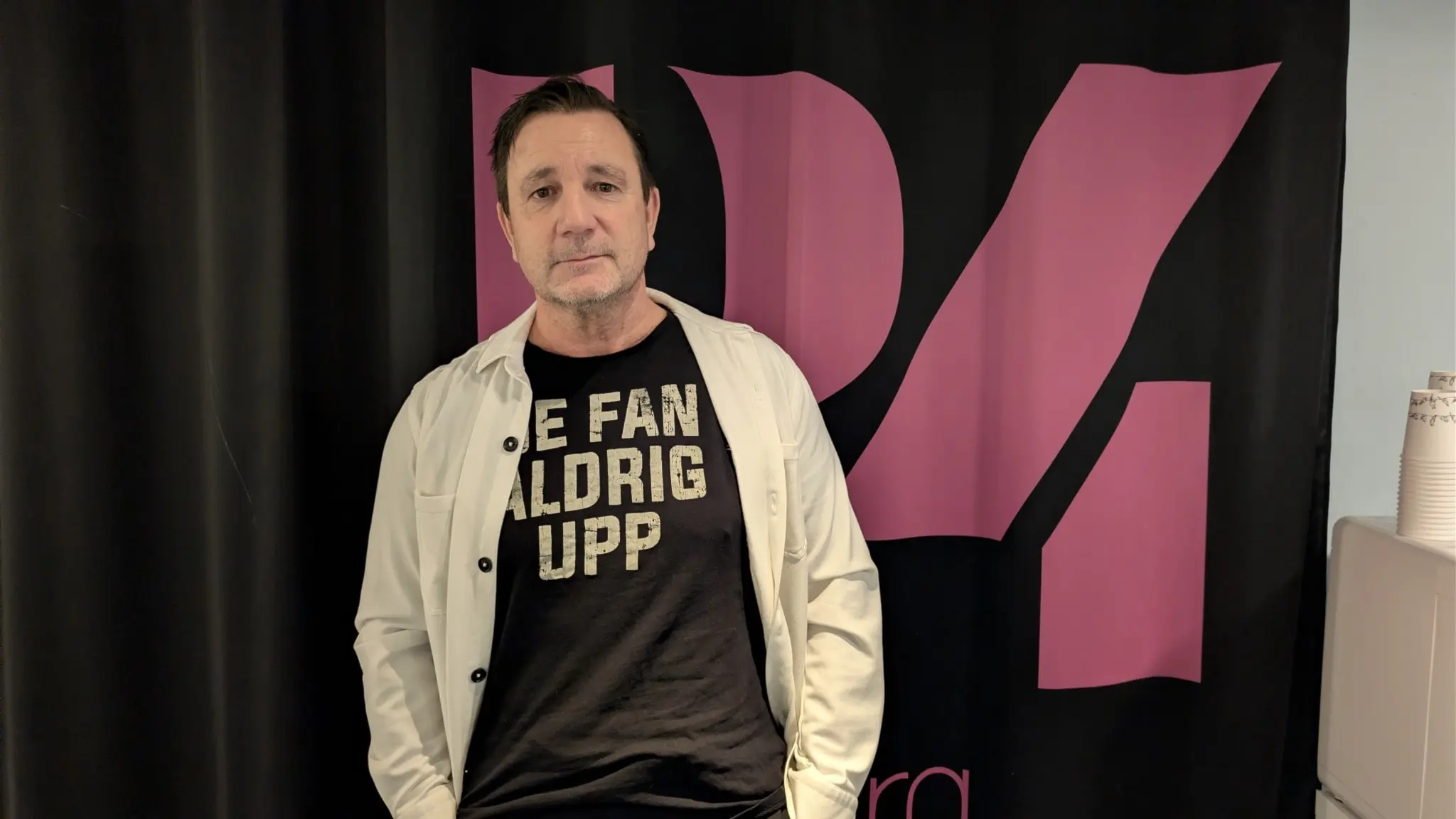 Peter Källberg från Gävle, efter sin canceroperation, står framför en svart och rosa bakgrund med texten 'Ge fan aldrig upp' på sin tröja. Han symboliserar styrka och kamp mot sjukdom.