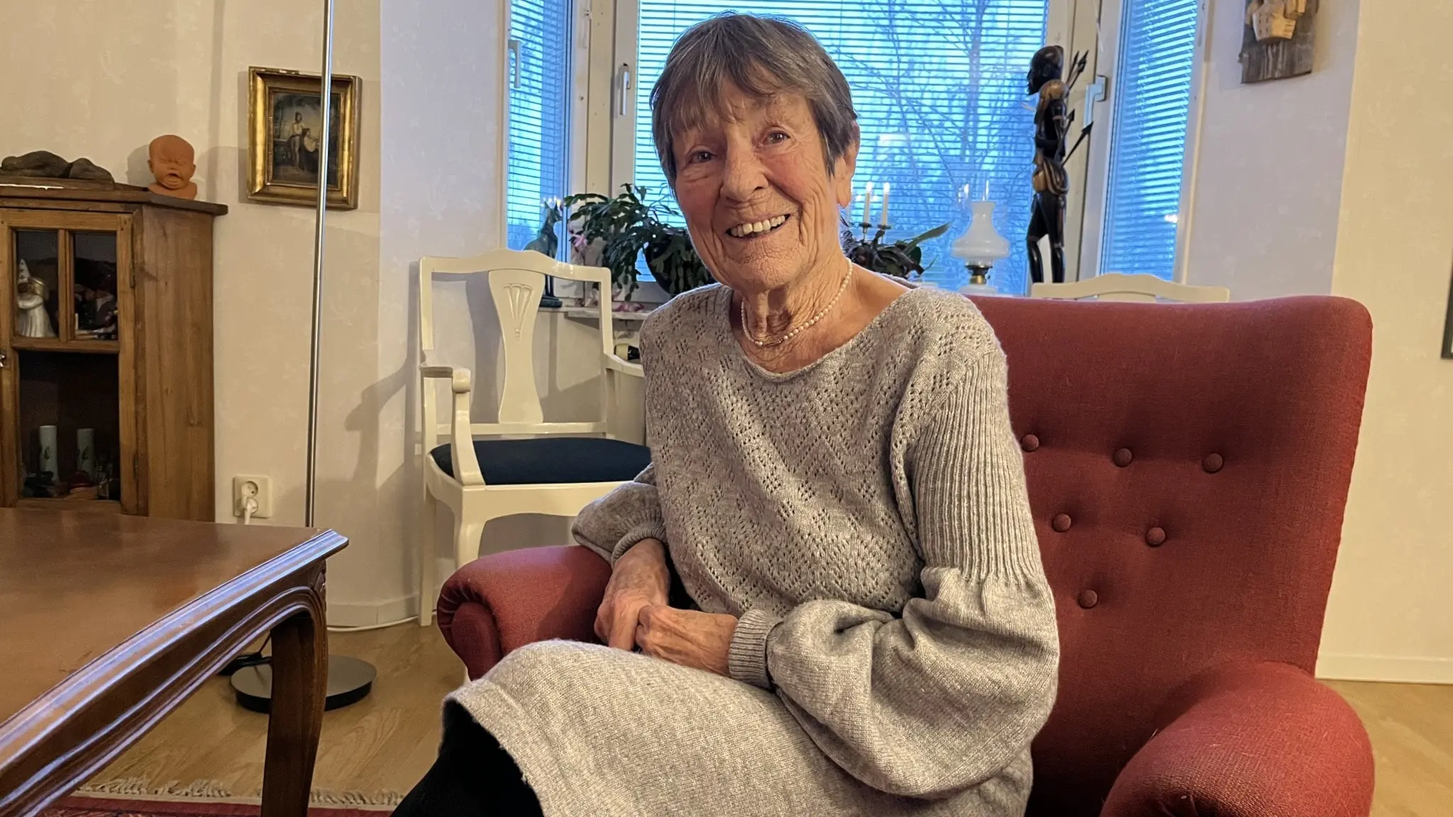 Kirstin Salin sitter i en röd fåtölj i ett vardagsrum med en stickad ljusgrå tröja och svarta byxor. I bakgrunden syns fönster med persienner och möbler.