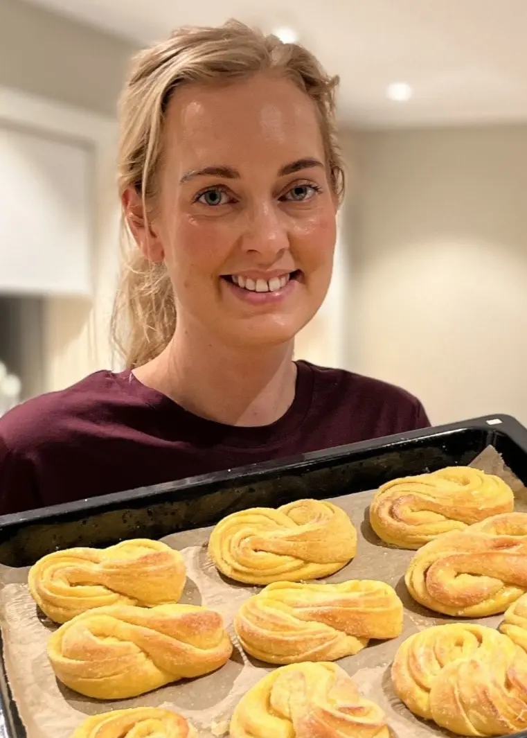 Mathilda Öman håller en plåt med nygräddade saffransbullar i ett modernt kök med ljusa väggar och diskbänk i bakgrunden.