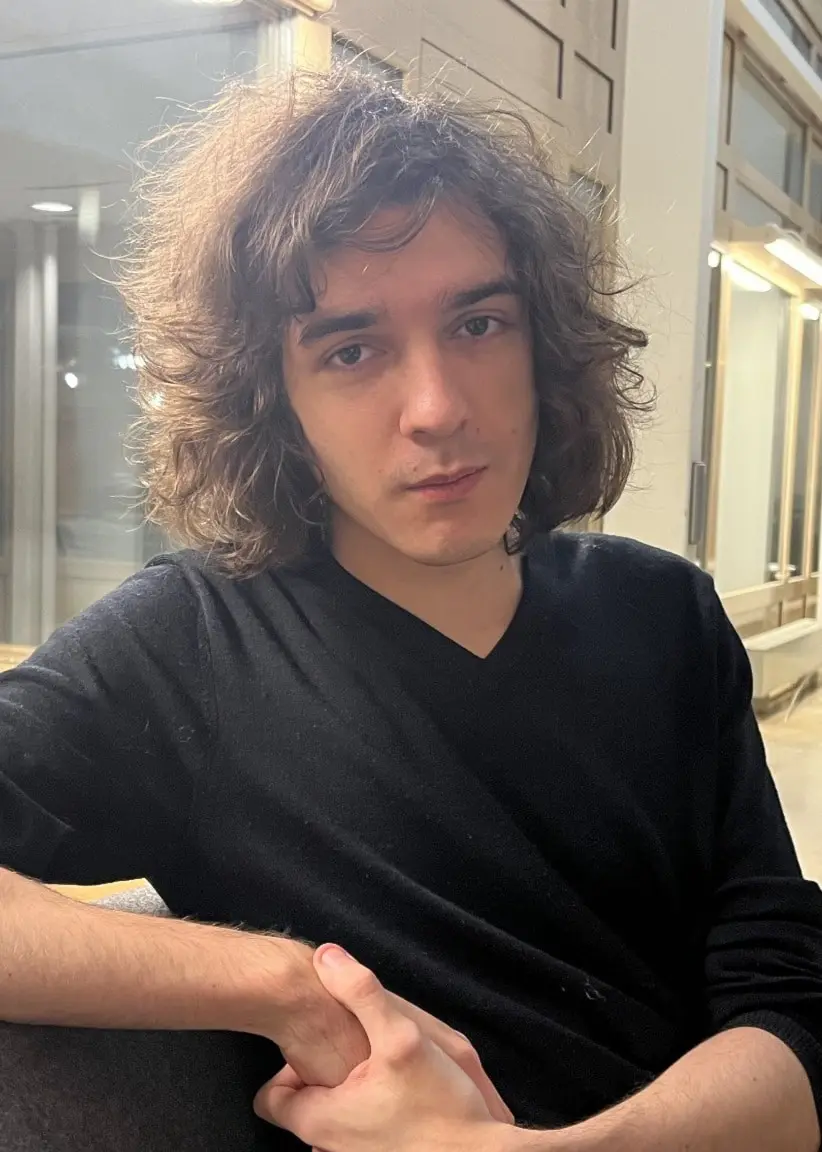 En ung kille med långt brunt och lockigt hår sitter avslappnat i en modern inomhusmiljö med stora fönster och ljusa väggar, iförd en svart tröja. Bakgrunden visar en öppen kontorsliknande miljö med möbler.