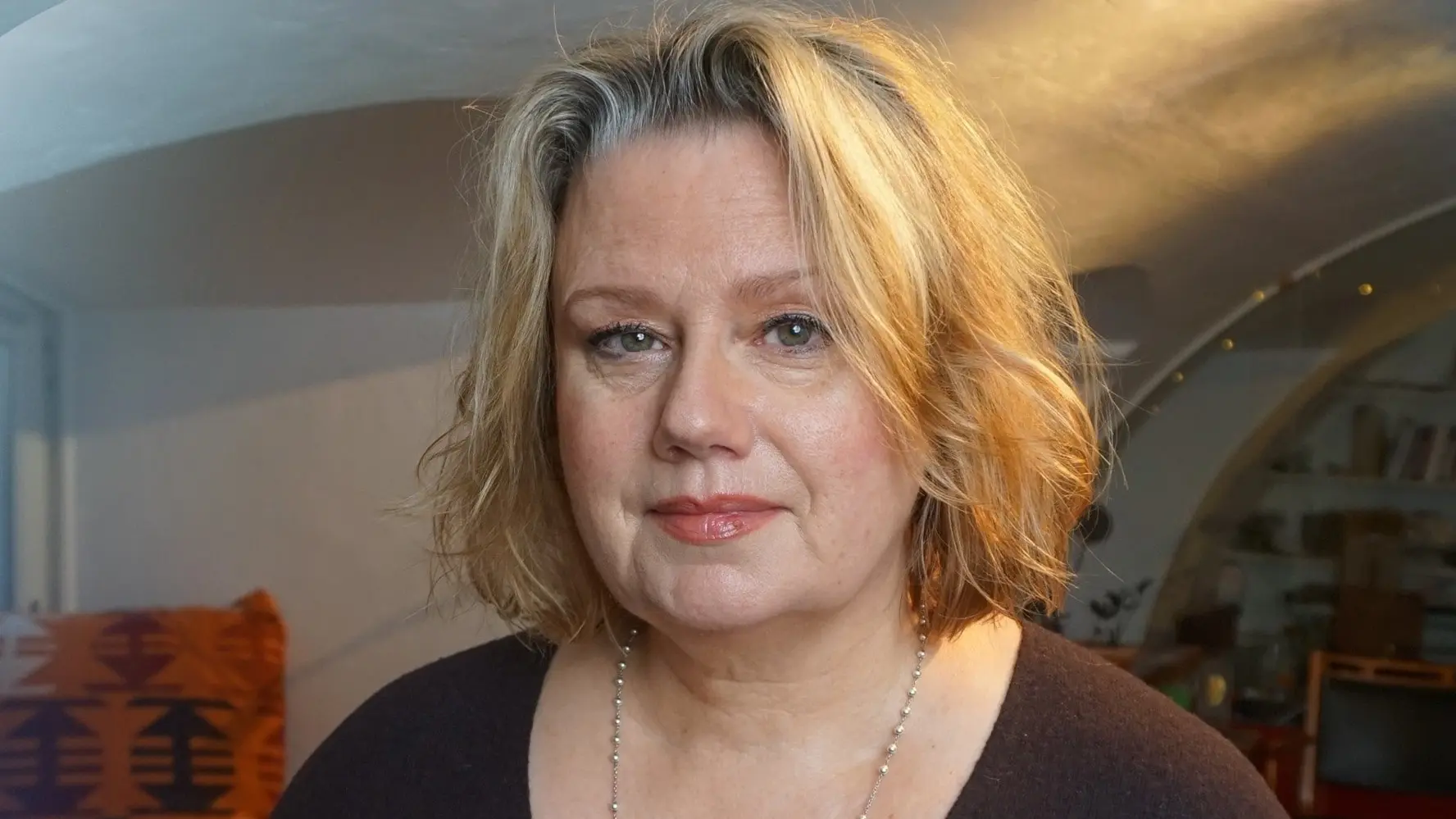 En kvinna med blont hår står inomhus i ett rum med välvt tak, bokhyllor och en orange kudde i bakgrunden. Hon bär en mörk tröja och ett halsband.