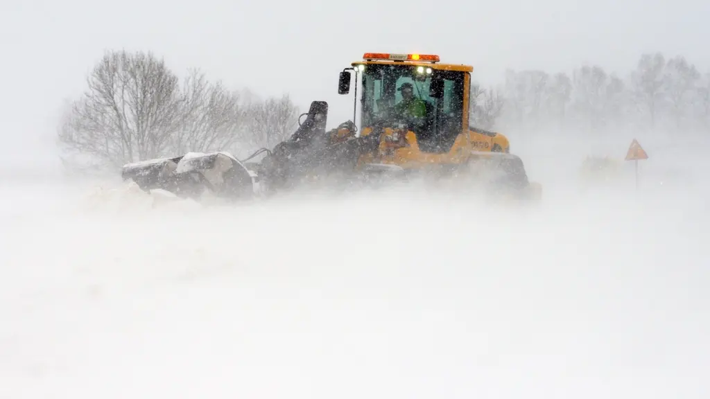 En snöplog arbetar för att röja vägen i kraftigt snöfall på Österlen. Maskinen är omgiven av tät snö och dimma, medan träd i bakgrunden skymtar genom det vita landskapet.