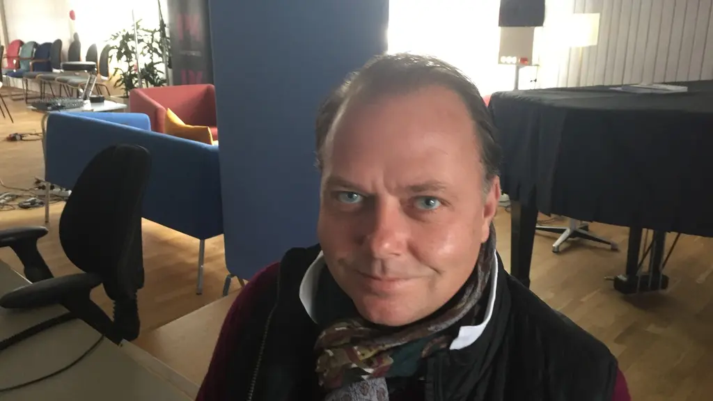 Man med scarve som tittar in i kameran. Han sitter i en studio.