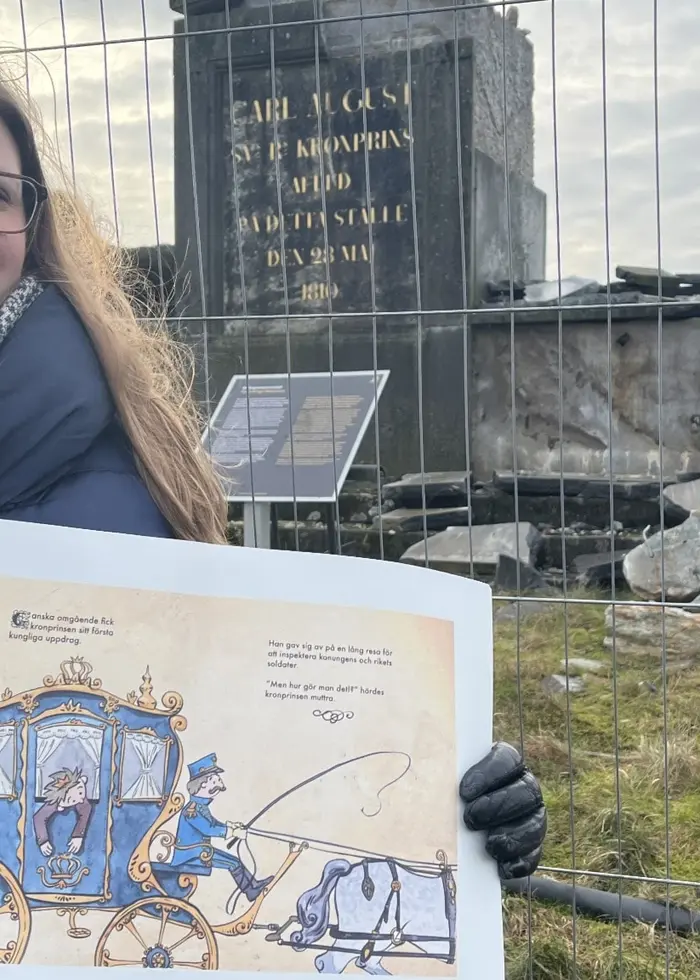 En person håller upp en illustration framför ett stängsel som omger ett område med ruiner och gräs. Bakgrunden visar en minnesplats med stenblock och skyltar. Himlen är molnig.