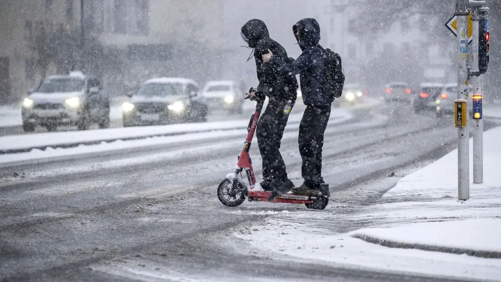 Två personer klädda i vinterkläder åker tillsammans på en elsparkcykel genom snöfall vid en trafiksignal i centrala Malmö. Bilar syns i bakgrunden och snön ligger på marken.