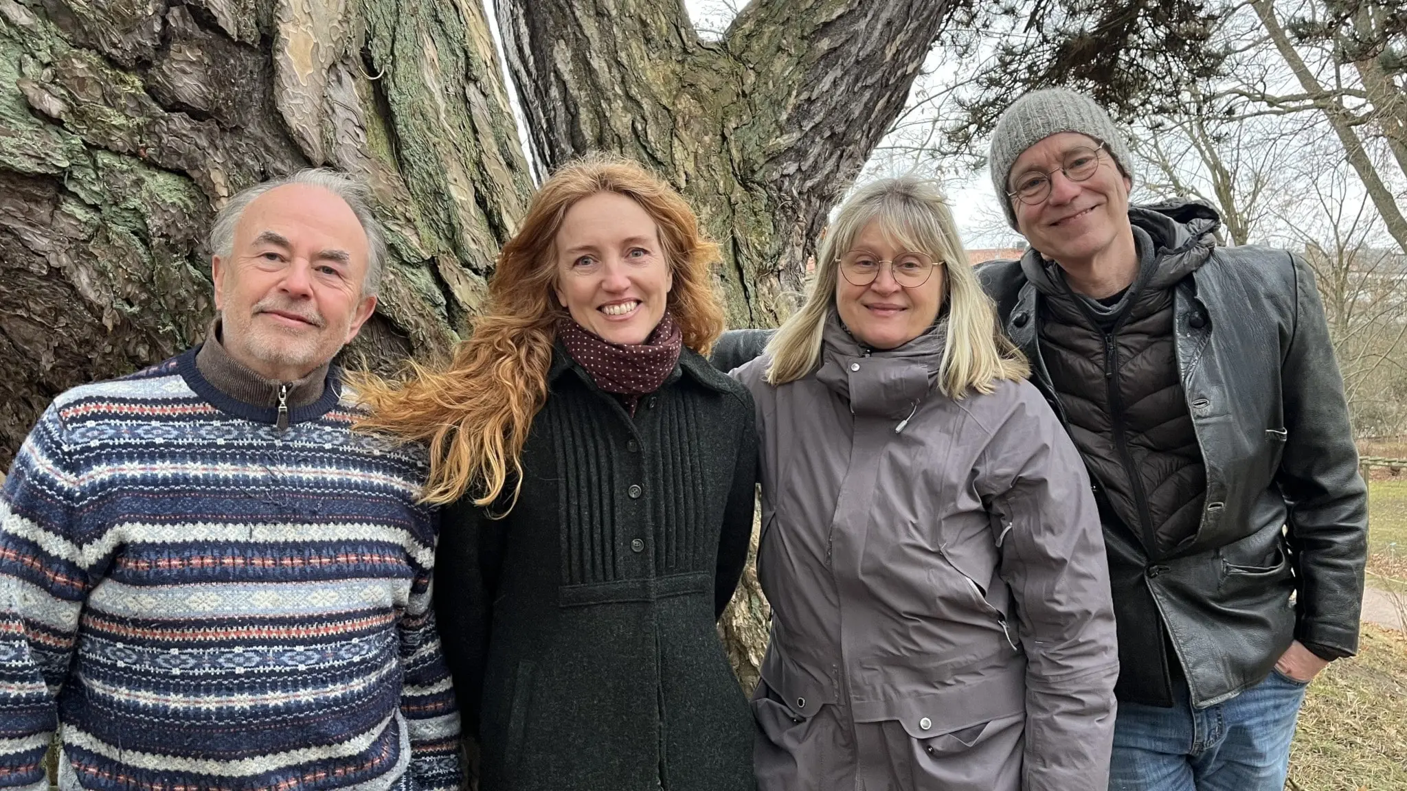 Fyra personer står utomhus i Botaniska trädgården i Lund framför ett stort träd. Två av dem har suddade ansikten, medan de andra två ler mot kameran. Alla är klädda för kyligt väder.