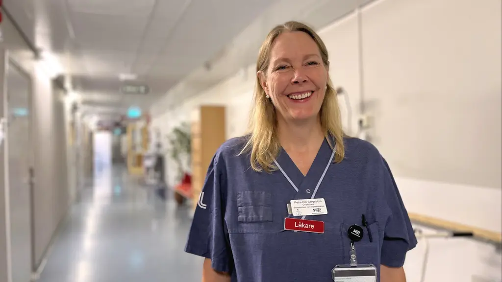 Barnläkaren Petra Um Bergström står i en sjukhuskorridor klädd i blå arbetskläder med namnbricka och ID-kort. Bakgrunden visar en lång korridor med medicinsk utrustning och dörrar.