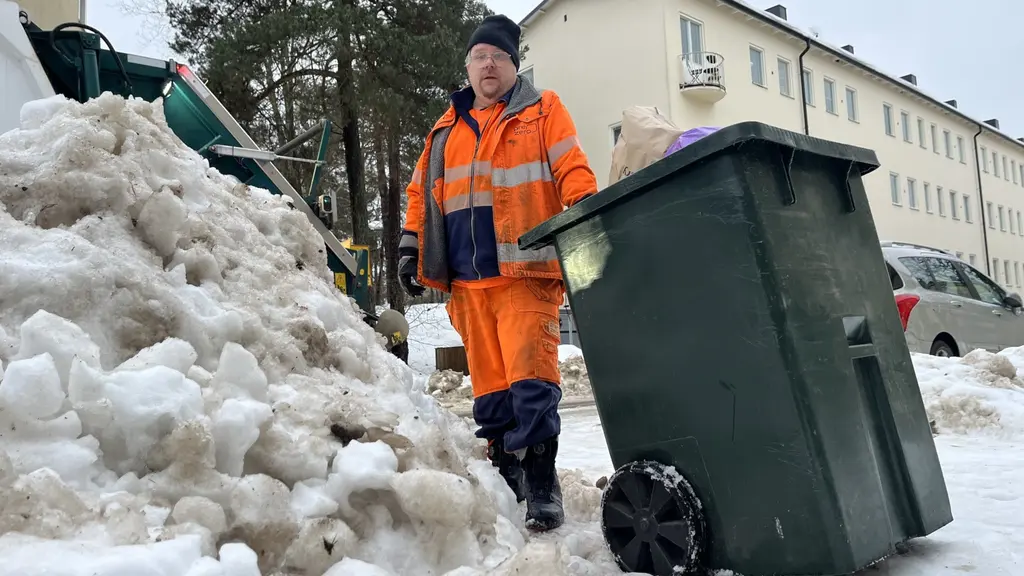 En person i orange arbetskläder drar en grön soptunna genom ett snöigt bostadsområde i Hammarbyhöjden. Snöhögar ligger vid sidan av vägen, och flerfamiljshus syns i bakgrunden.
