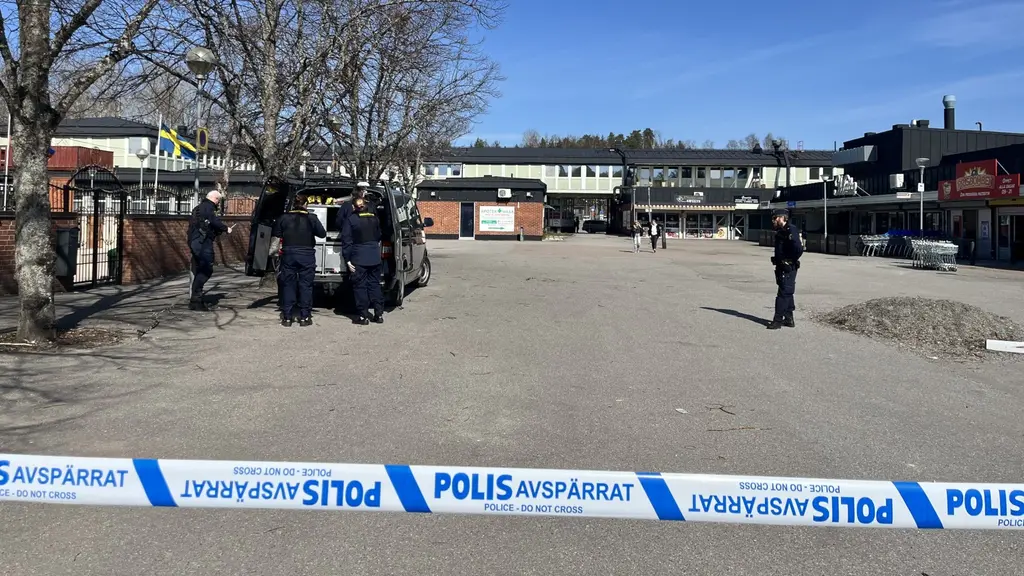 En avspärrad plats utomhus med polistejp och flera poliser på plats. Området är tomt och omges av byggnader samt några träd. Himlen är klarblå.
