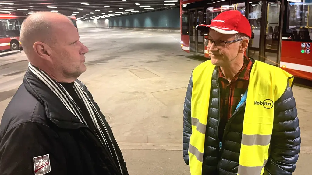 Två män, Peter Sellén och Nils Sjöblom, talar med varann på en busstation, den ene klädd  i kavaj, den andra i reflexväst.