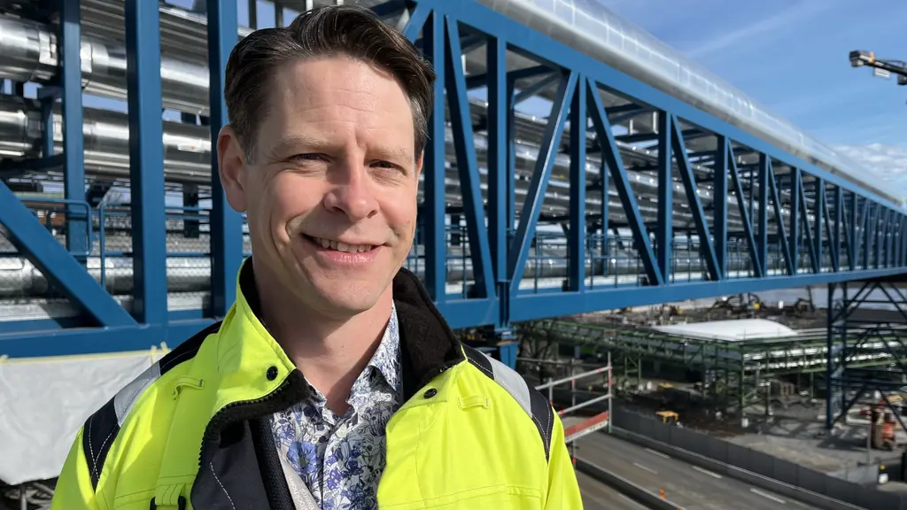 En person i gul reflexjacka står utomhus vid en stor byggnadsstruktur av stål och glas. Bakgrunden visar en modern bro eller gångväg som sträcker sig över området.