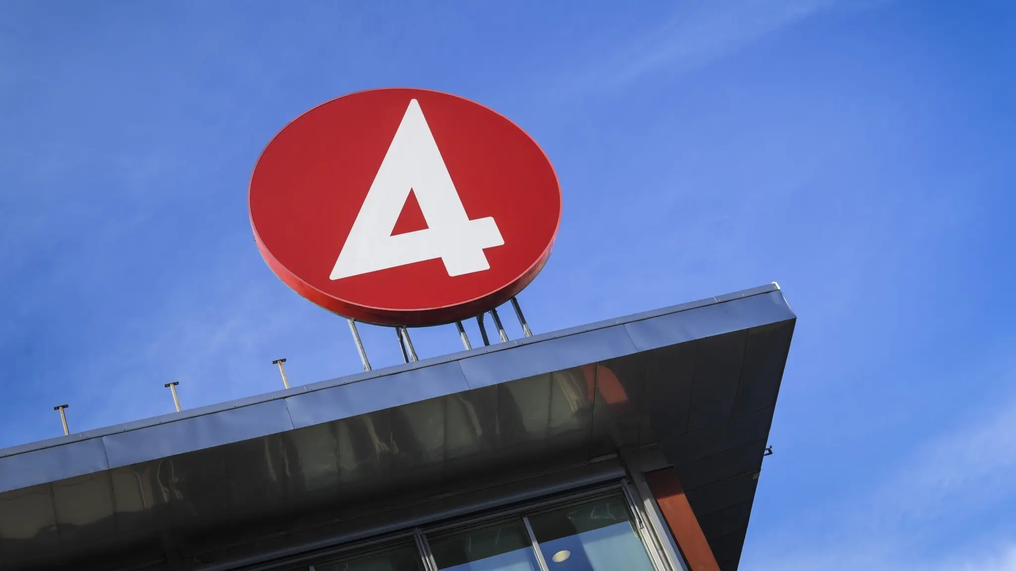 The image shows a large, red, four-sided sign with a white "4" prominently displayed against a clear blue sky. The sign is mounted on a metal structure that extends from the top of a building, which has a dark gray or black roof. The building's facade is primarily gray or dark gray, with a section of reddish-brown or b