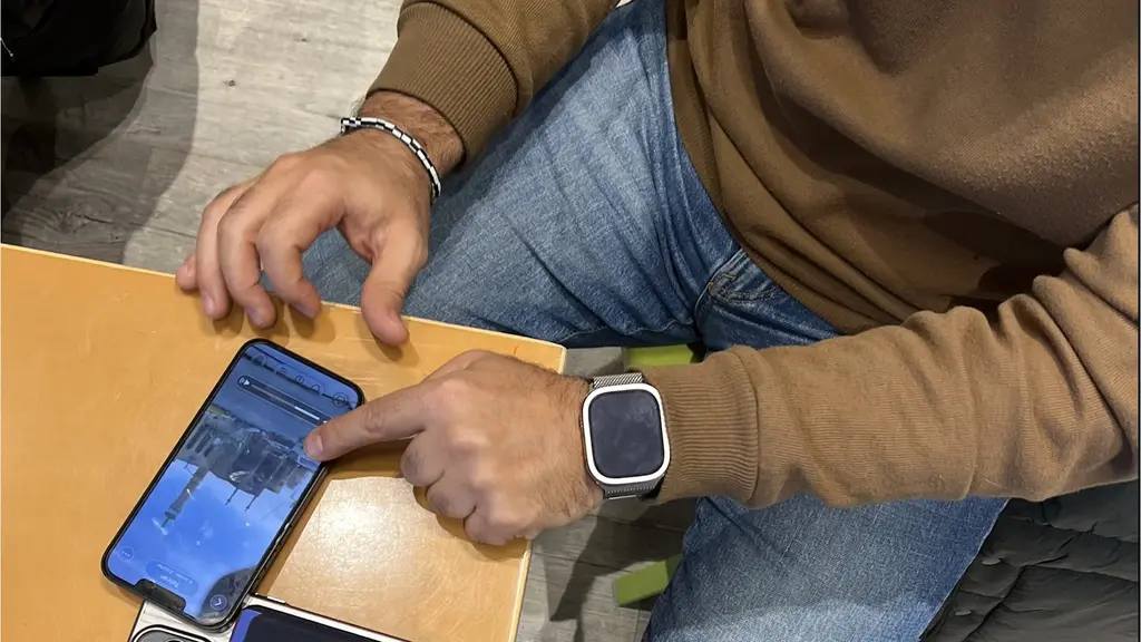 En person sitter vid ett bord och pekar på en smartphone med en bild på skärmen. Personen bär en brun tröja, blå jeans och en smartklocka. En annan mobiltelefon ligger på bordet.