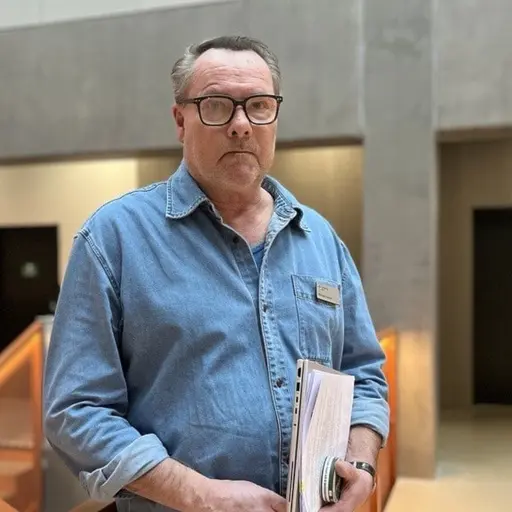 En person i jeansskjorta står inomhus i en modern byggnad med betongväggar och trädetaljer. Personen håller papper i händerna och bakom syns trappor och dörrar.