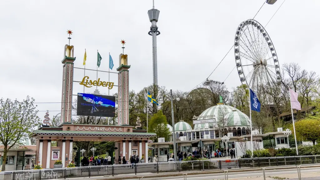 Norra entrén till Liseberg i Göteborg med färgglada flaggor, nöjesparkens skylt, en stor pariserhjul och utsiktstorn i bakgrunden. Människor rör sig vid entrén på en mulen dag.