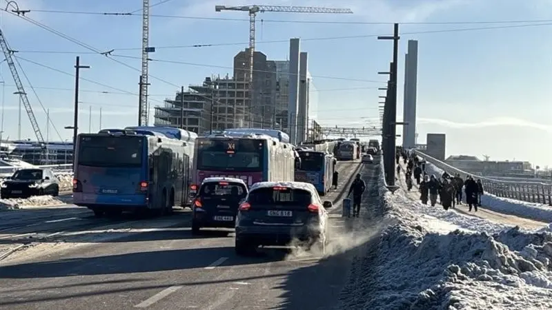 En solig vintermorgon på Hisingen med snö på marken. Bussar, bilar och fotgängare delar en bro, omgivna av byggkranar och höga byggnader i bakgrunden.