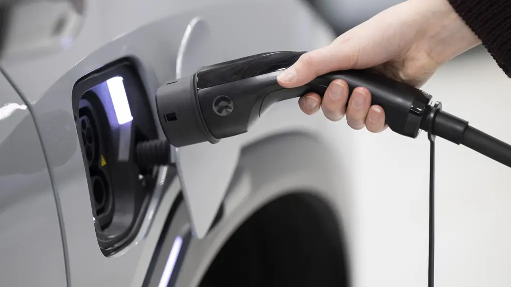 En närbild visar en hand som ansluter en laddkabel till en elbil. Bilen är silverfärgad och laddningsporten lyser med ett blått sken, vilket indikerar att laddning pågår. Fokus ligger på hållbarhet och teknologi.