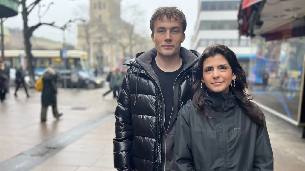 Två personer står på en trottoar i en stadsmiljö. Den ena bär en svart dunjacka och den andra en mörk jacka. I bakgrunden syns en spårvagn, människor som promenerar och stadens byggnader.