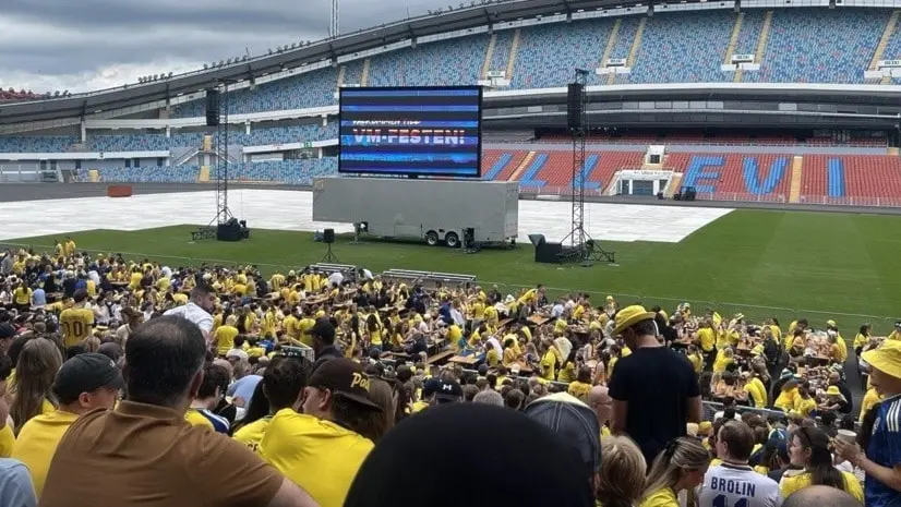 Fullsatt läktare på Ullevi med Sverige-supportrar i gult framför storbildsskärm på planen, under mörka moln inför landskampen där Sverige kraschade.