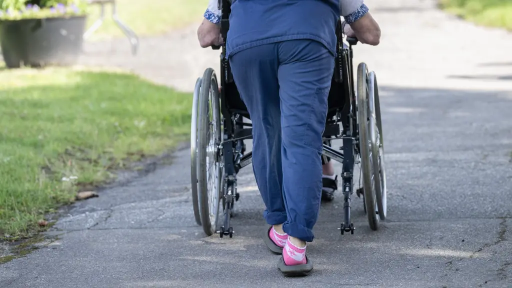 En person i arbetskläder skjuter en person i en rullstol framför sig på en gångväg i solen.