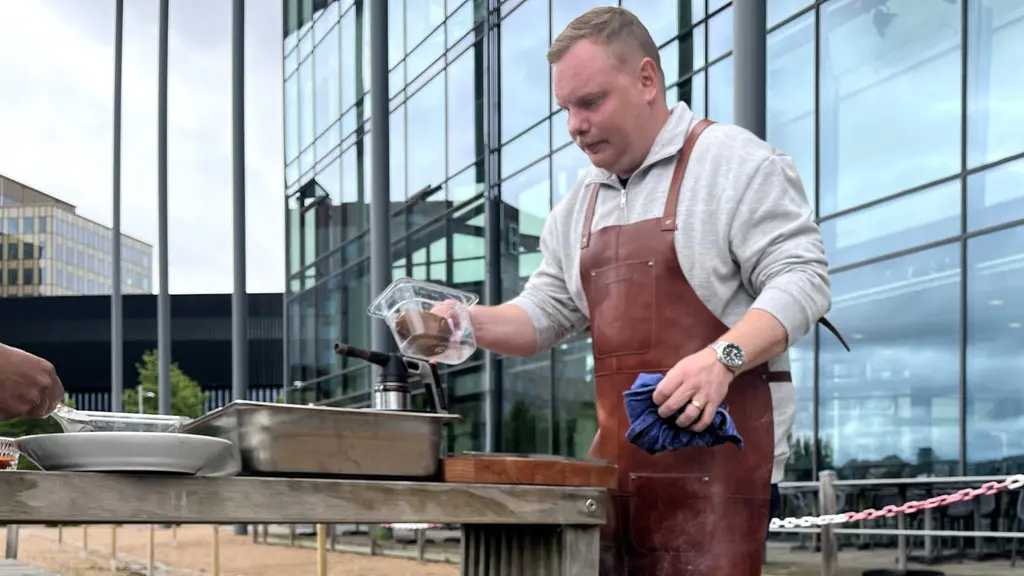 Alexander Stärnerz grillar vid en utomhusstation utanför modern glasbyggnad, med rykande kolgrill och matlagning i fokus.