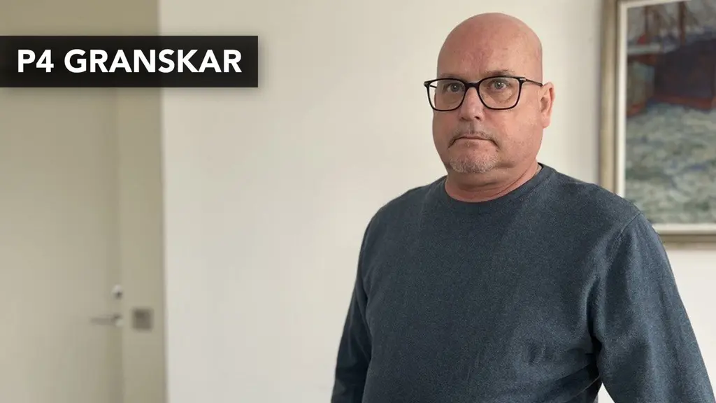 En person i grå tröja står inomhus framför en vit vägg med en dörr till vänster och en tavla till höger.