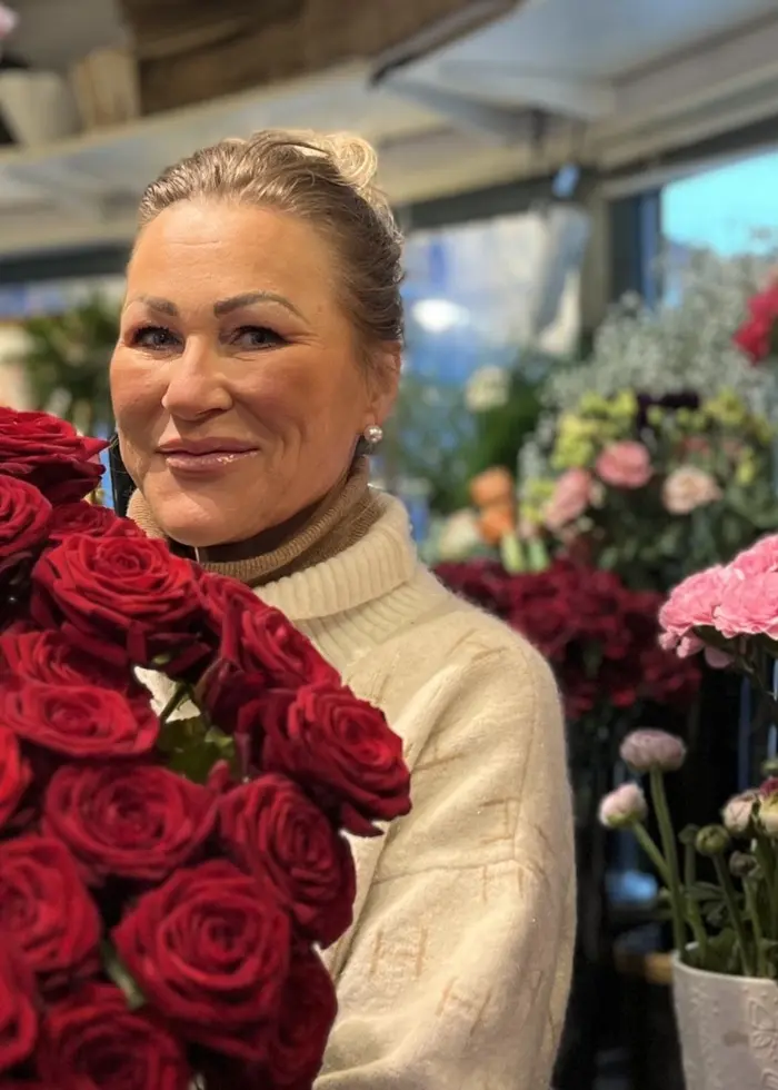En blomsterbutik fylld med röda rosor och rosa blommor. En person i ljus tröja håller en stor bukett röda rosor. Bakgrunden visar fler blommor och ett skyltfönster med dagsljus.