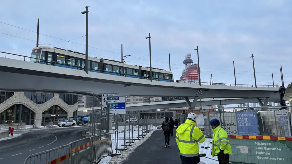 Hisingsbron i Göteborg visas med avspärrningar och två personer i gula reflexjackor som diskuterar vid en avspärrad väg. En stillastående spårvagn syns ovanpå bron, och himlen är molnig.