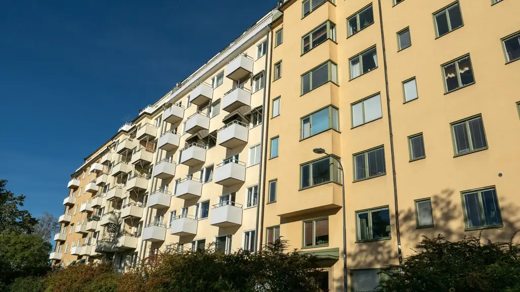 Bostadshus med ljus fasad och balkonger. Soligt väder och klarblå himmel. Grönska syns framför byggnaden.