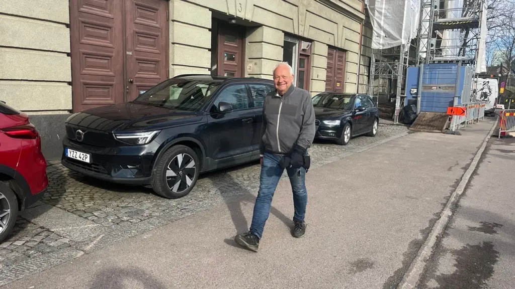 Johan Gunnebo klädd i grå jacka och blå jeans promenerar längs en trottoar. Intill honom står flera parkerade bilar, inklusive en mörkblå SUV. I bakgrunden syns en byggnad med trädörrar och en byggarbetsplats.