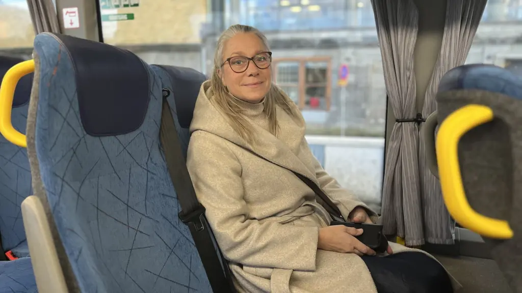 Tove Berggren sitter i en buss iklädd beige kappa, med en svart väska över axeln. Bussens inredning har blå säten och gula handtag. Utanför syns en byggnad med glasfasad.