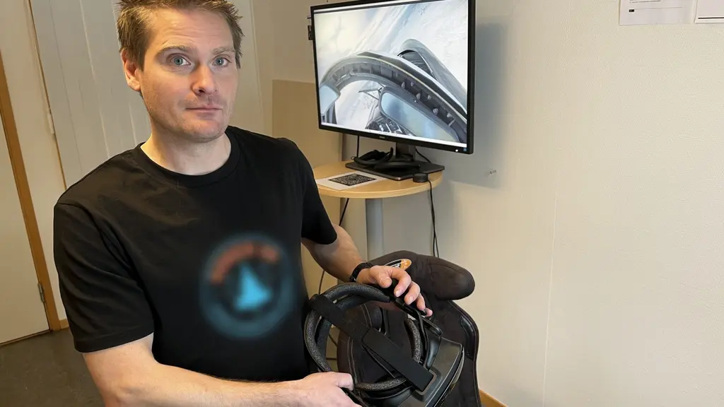 En person håller en VR-utrustning och står i ett rum med en TV som visar en bild relaterad till VR. Personen bär en svart t-shirt med logotypen för hans kanal.