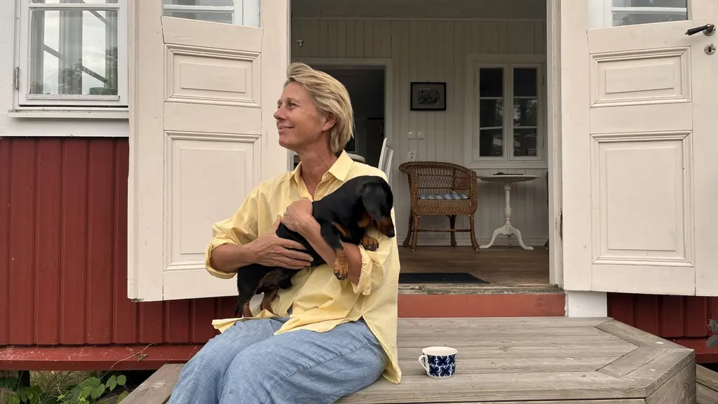Lina Serning sitter på farstukvisten på Tjurkö med en tax i famnen, framför ett rött trähus med öppna vita dörrar. Hon bär en gul skjorta och blå jeans.