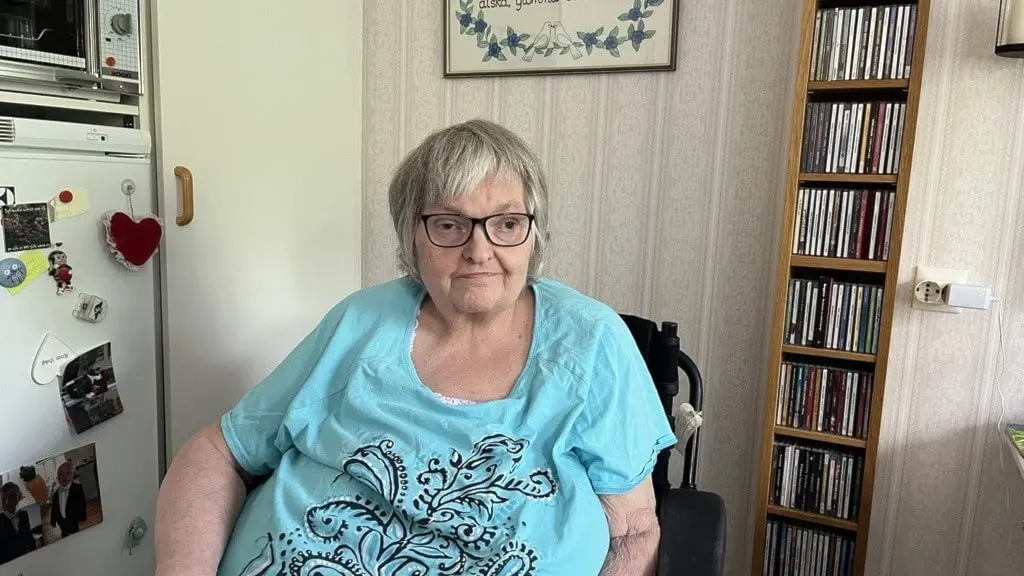 Porträtt av Ann-Sofie Pamp i rullstol hemma, sittande vid köksbordet i ljusblå tröja. Kylskåp och CD-hylla syns i bakgrunden.