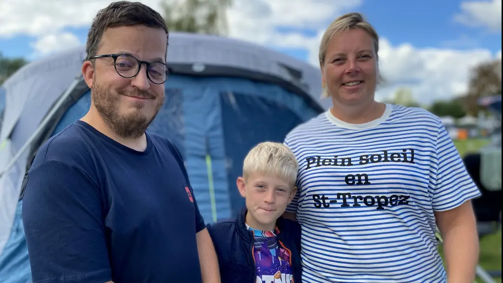 En familj står framför ett blått tält på en campingplats. En vuxen man, en vuxen kvinna och ett barn ses tillsammans i bilden. Himlen är blå med vita moln i bakgrunden.