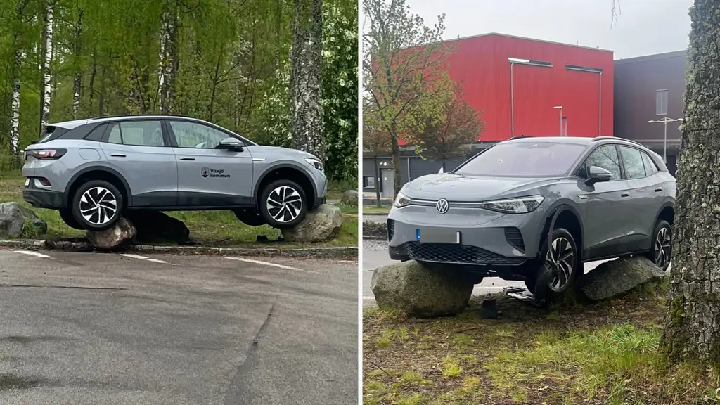 En grå tjänstebil från Växjö kommun har kört upp på stora stenar. Bilen står med hjulen på olika nivåer, vilket skapar en lutande position. Bakgrunden visar en röd byggnad och grönskande träd.