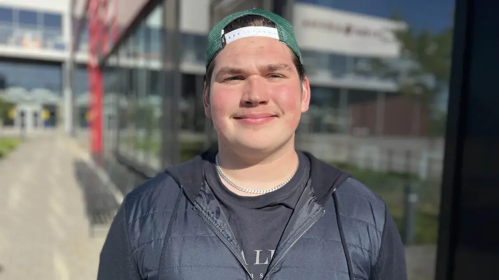 Edvin Nilsson står utomhus framför en modern byggnad med glasfasad. Solen skiner och det är klarblå himmel, vilket ger en ljus och skarp kontrast i bilden.