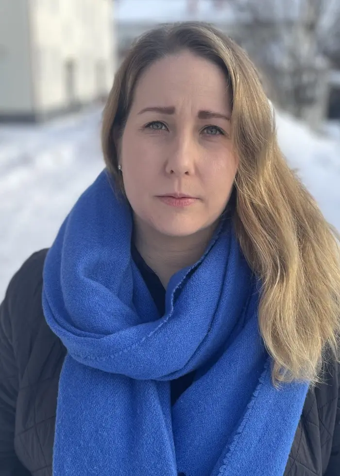 Meiju Vartiainen är klädd i svart jacka och blå halsduk. I bakgrunden syns tegelhus och träd täckta av snö.