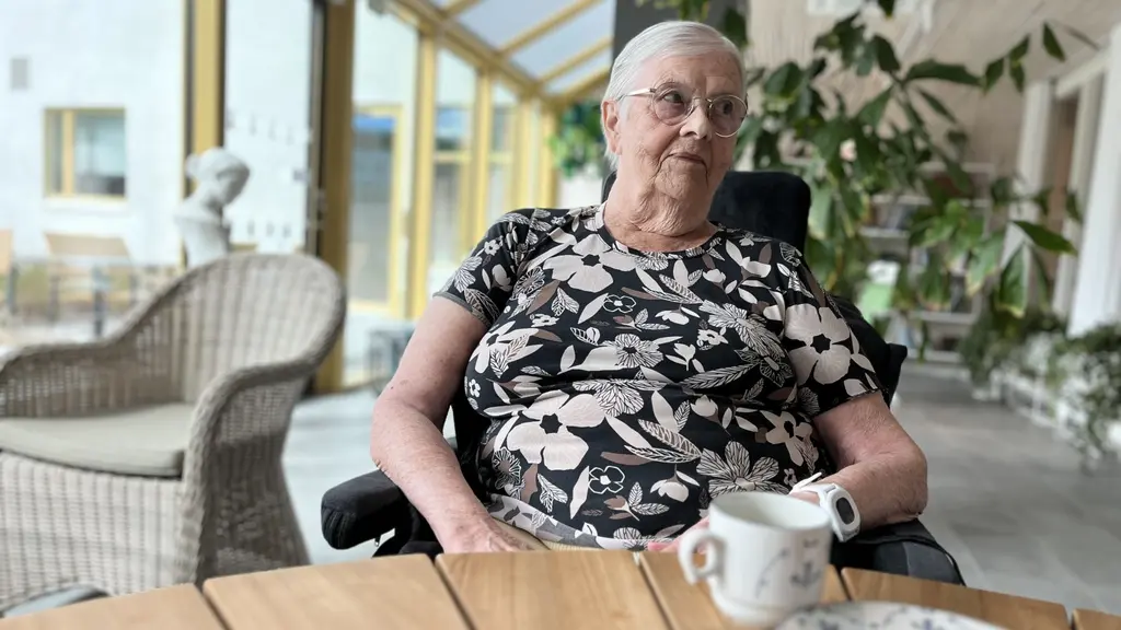 Ingrid Eriksson sitter vid ett träbord med en kopp kaffe. I bakgrunden syns växter, en glasvägg och utemöbler.
