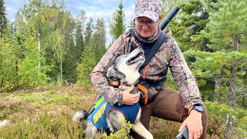 Gerd Lövgren sitter i skogsmiljö med hunden Tikka, som bär en blå väst med gul markering. Gerd har jaktkläder och en bössa över axeln, omgiven av grönskande träd och mossa.